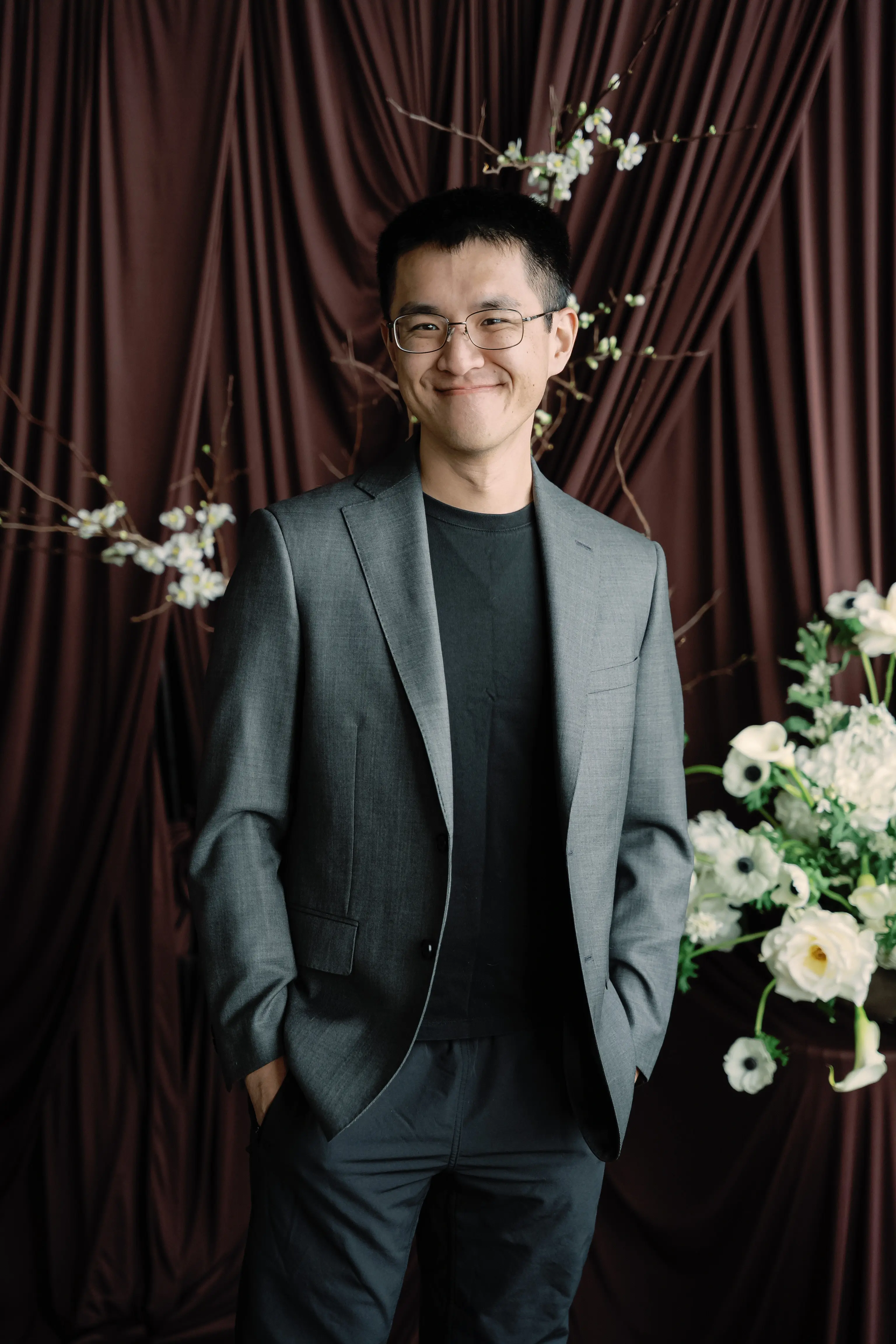 Gavin in a grey suit and glasses, hands in his pockets, standing between white anemones and blossom branches against a dark brown curtain
