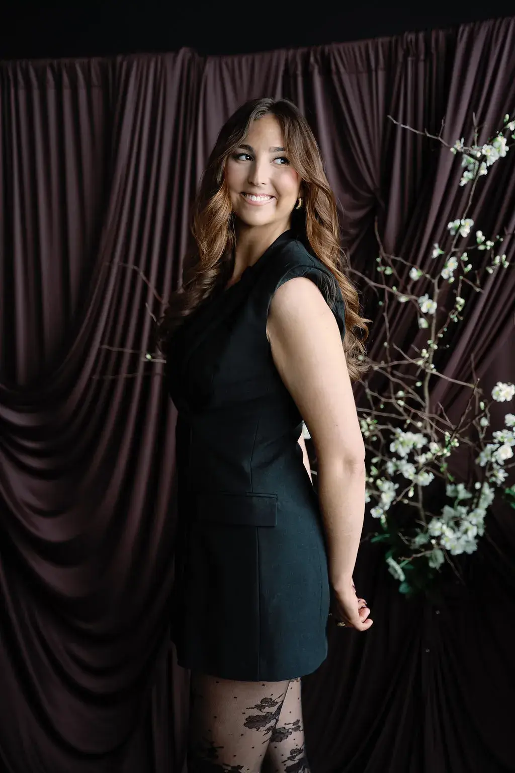 Jorja standing in profile in a black sleeveless dress and floral tights, long auburn hair over one shoulder, beside white blossom branches against a dark brown curtain