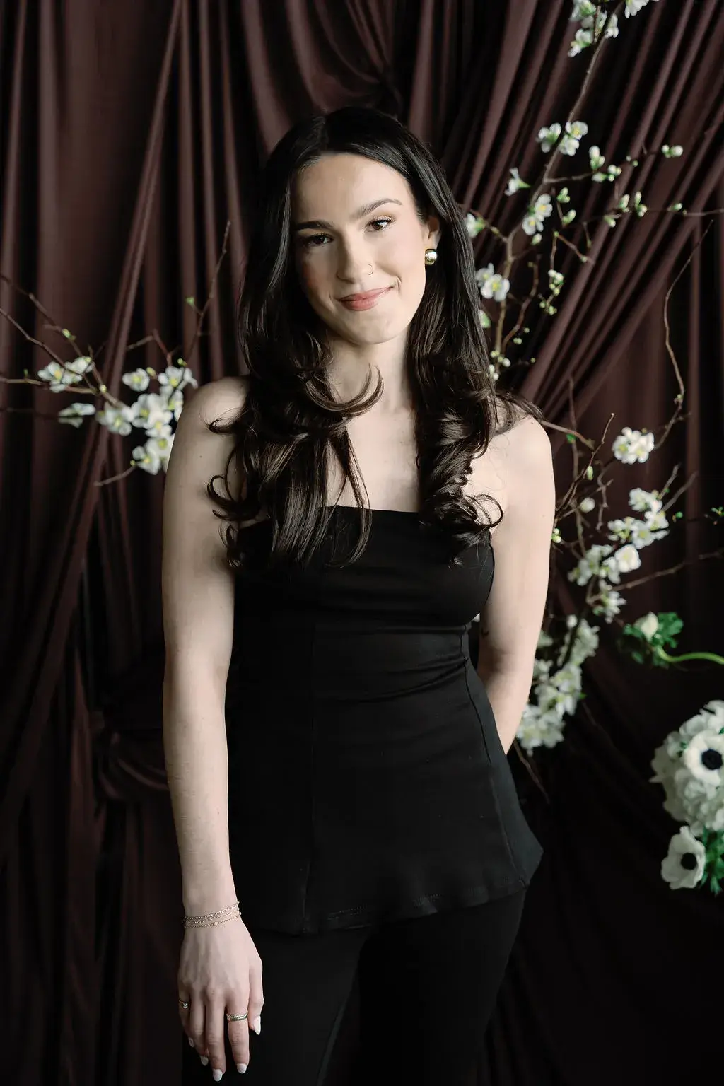 Maia in a black sleeveless jumpsuit, long dark waves falling past her shoulders, white blossom branches behind her against a dark brown curtain