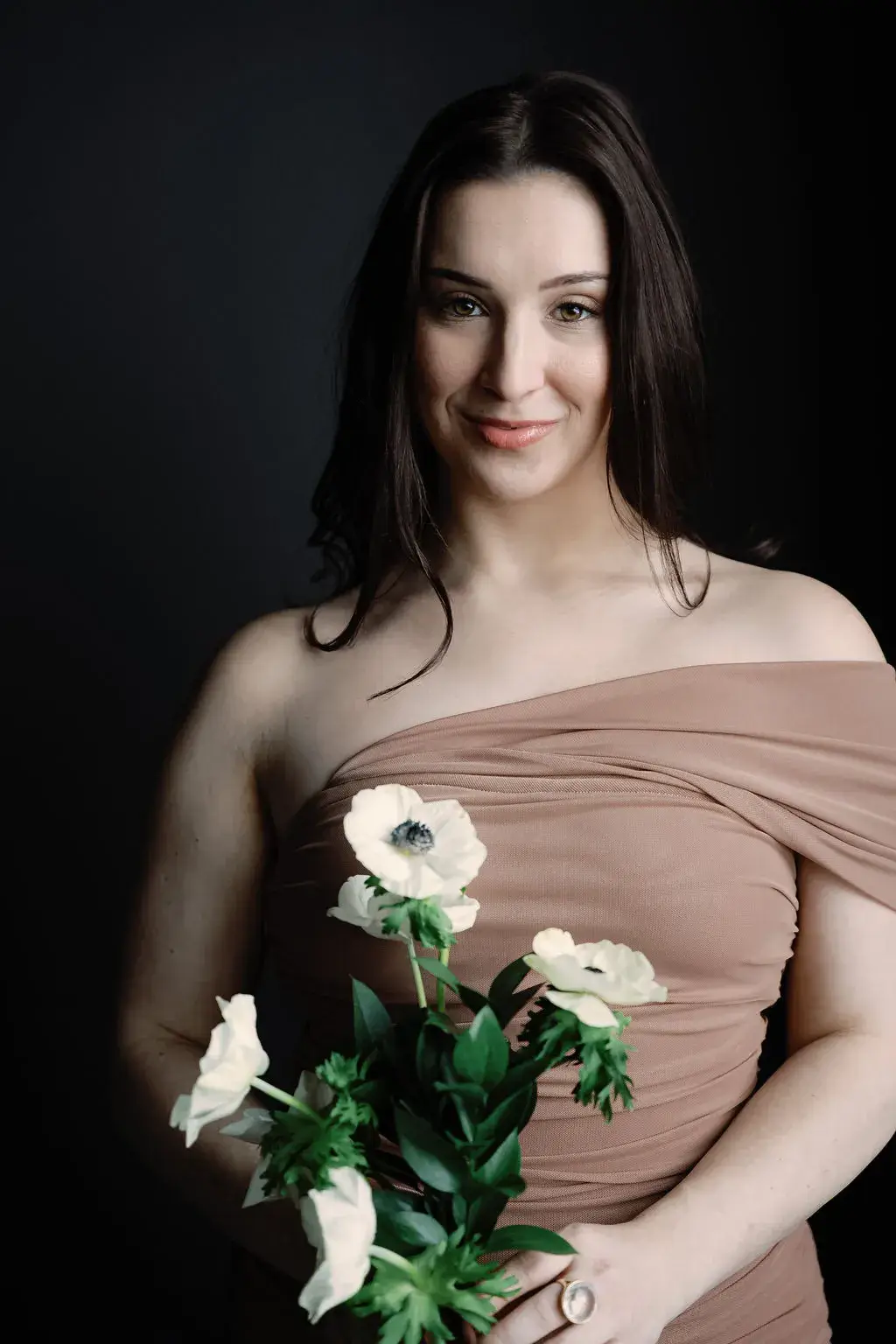 Shera in an off-the-shoulder blush gown, dark hair swept back, holding white calla lilies and anemones against a black backdrop