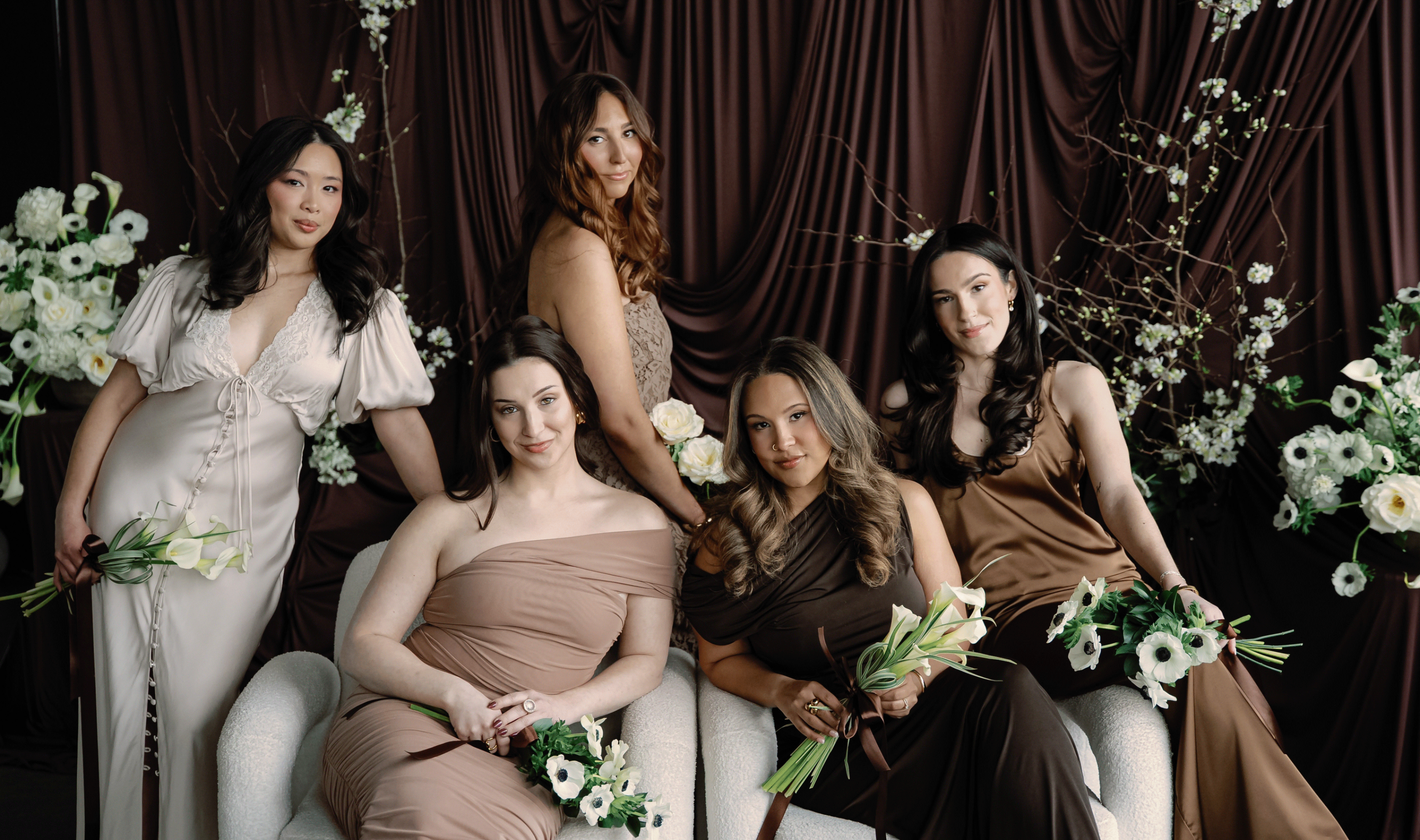 The five women of the Vivid Moments with San team in neutral silk and lace gowns, seated and standing with white anemones and calla lilies against a deep brown curtain
