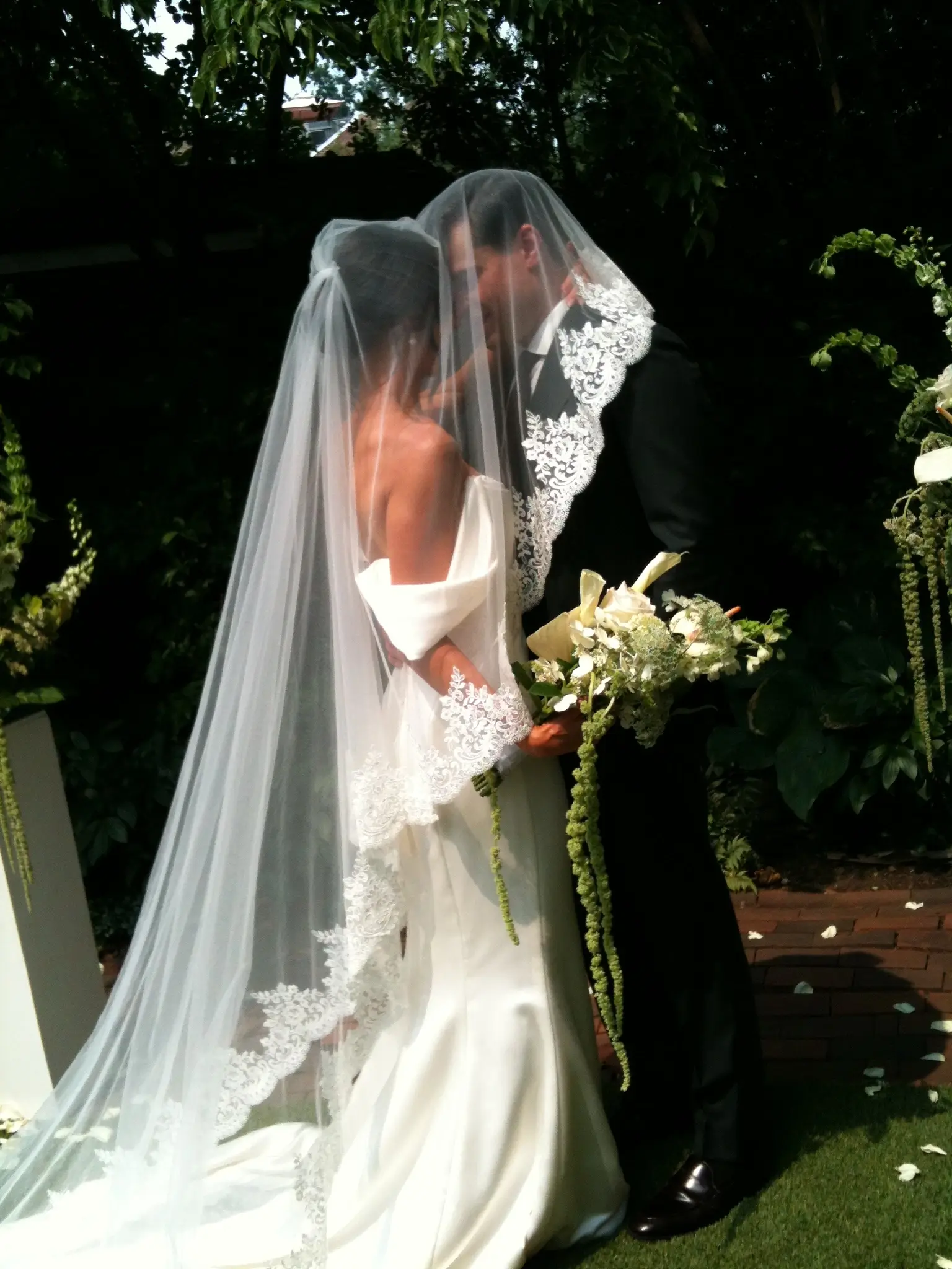A bride and groom kissing beneath a lace-edged cathedral veil in a garden, her bouquet of white calla lilies and trailing green amaranthus at her side