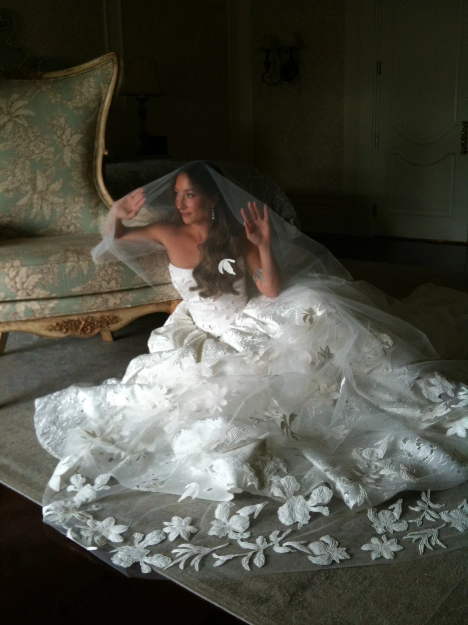 A bride sitting on the floor beside an antique settee, holding her cathedral veil out over the embroidered floral train of her gown