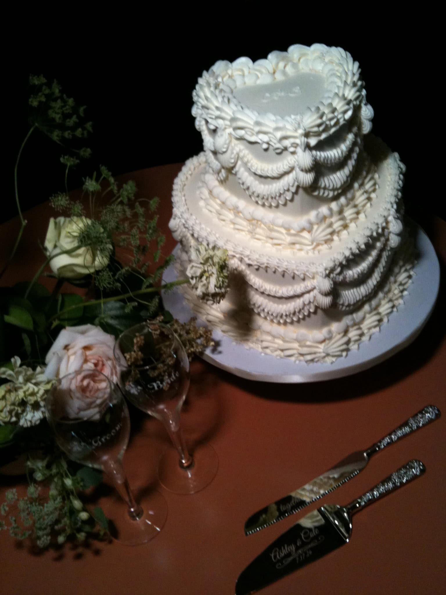 A tiered ivory buttercream wedding cake piped with ruffles and scallops, set beside garden roses, engraved champagne flutes and a cake knife
