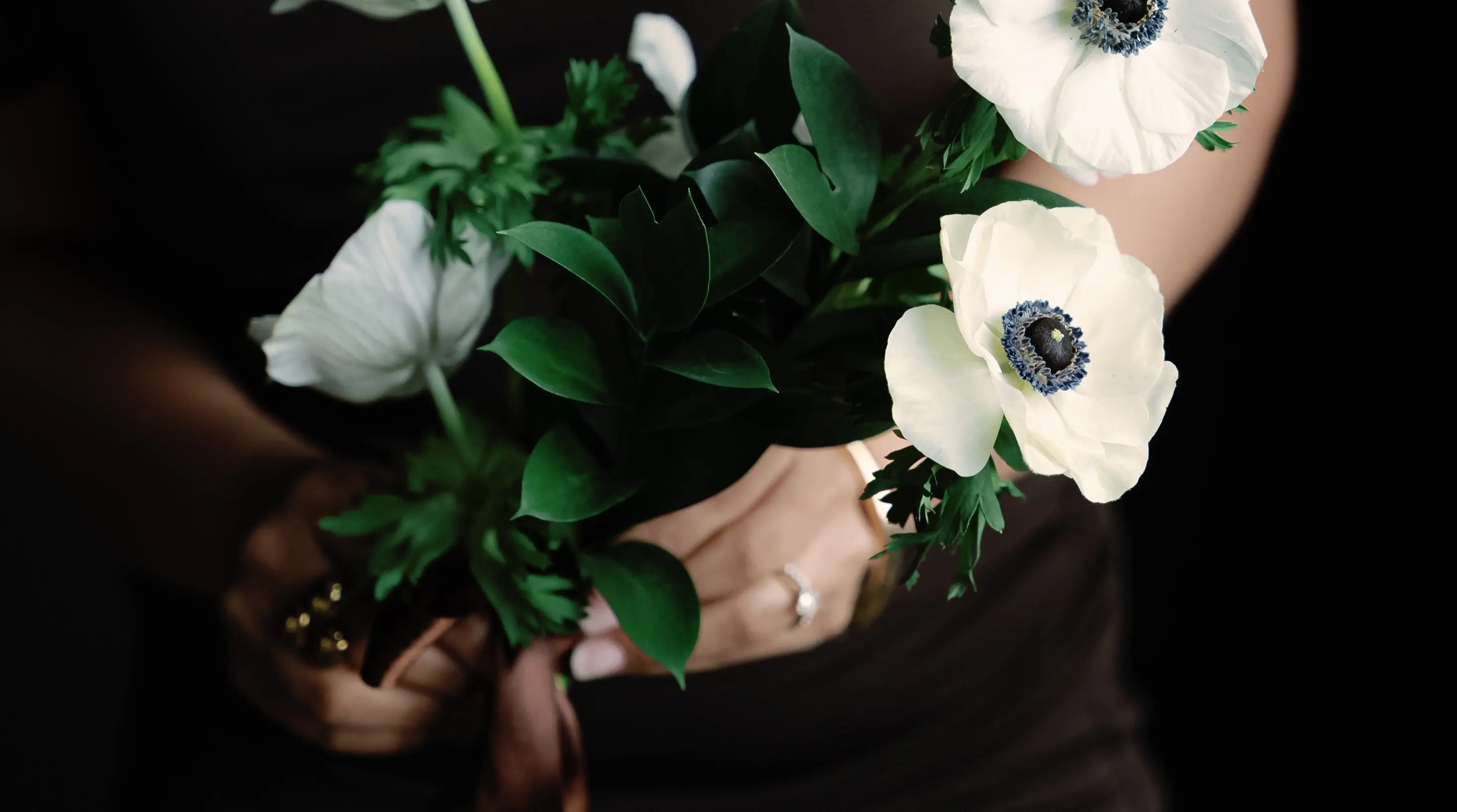A bouquet of white anemones and dark greenery held against a deep brown dress