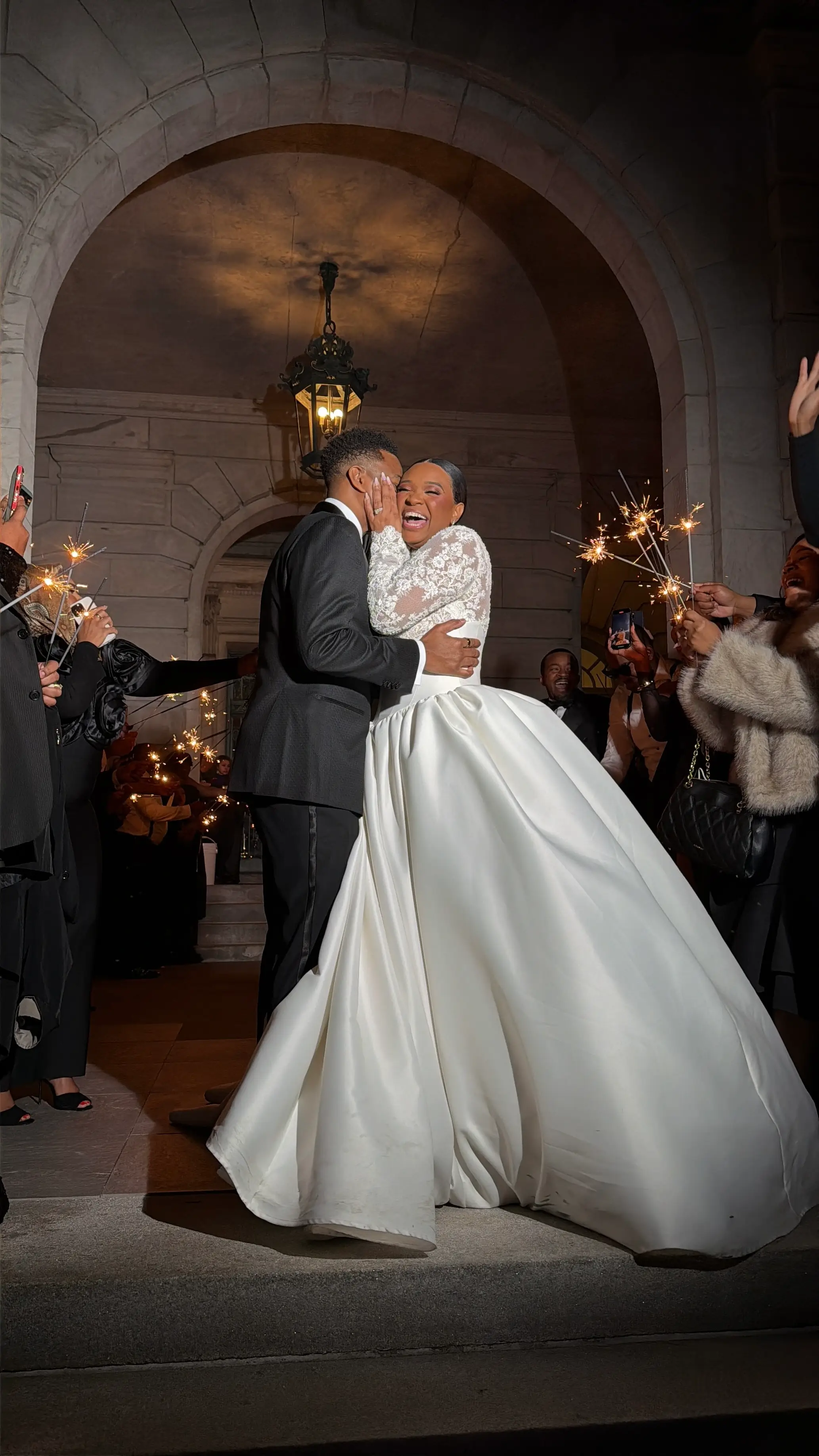 A couple embracing beneath a stone archway while guests hold sparklers around them