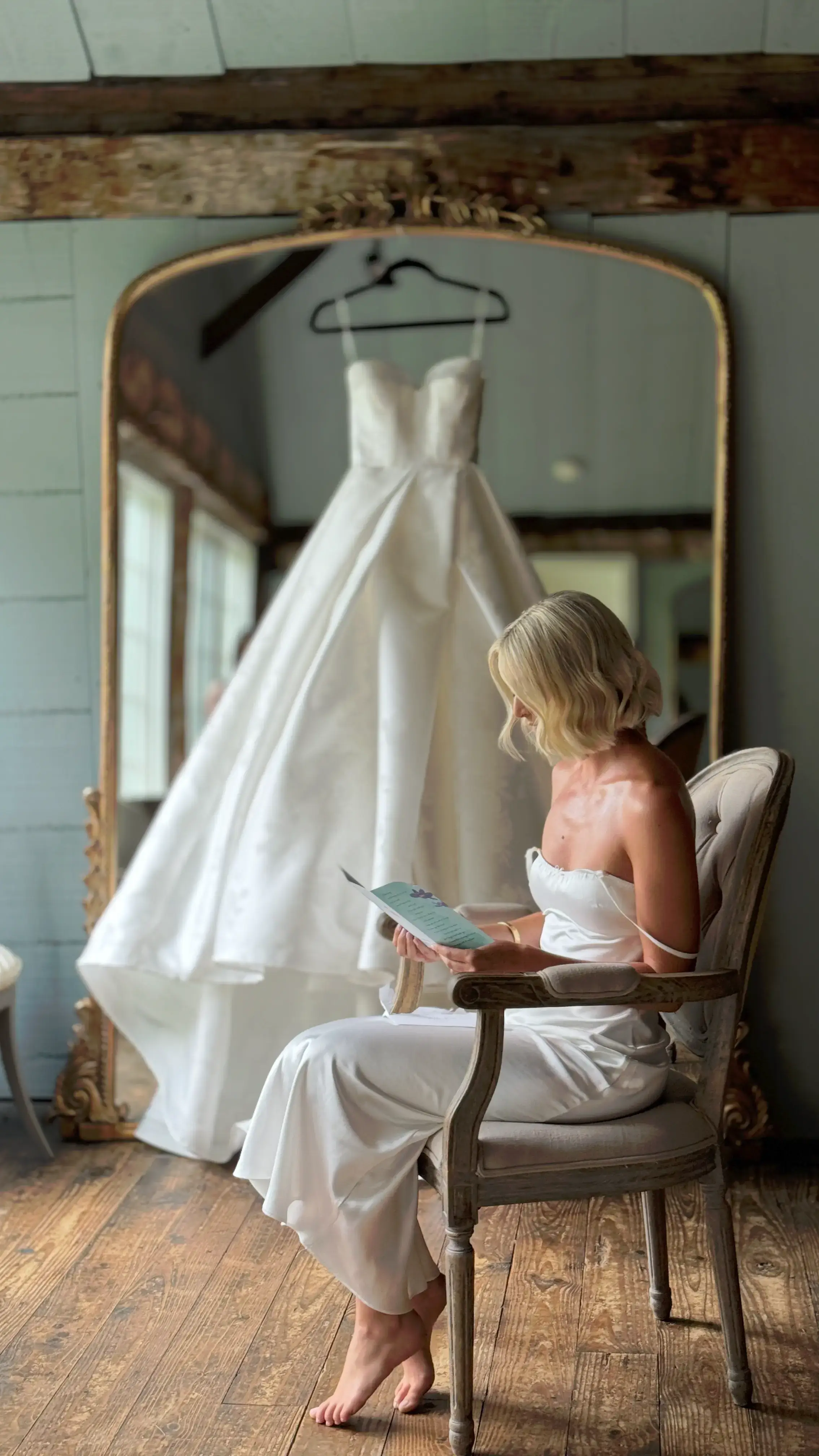 A bride in a slip dress seated beside her wedding gown hanging in front of a gilded mirror